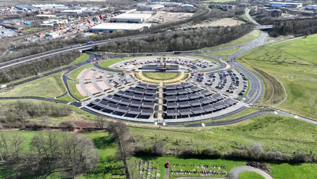 Aerial view of a large circular car park fitted with multiple rows of solar carport canopies surrounded by roads, green spaces, and industrial buildings.