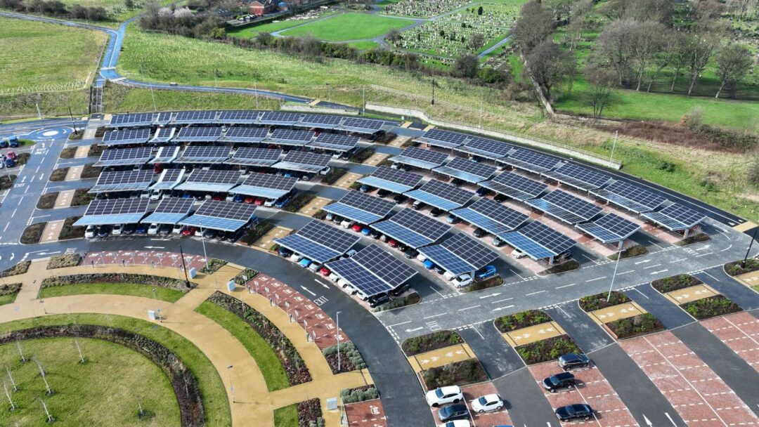 Aerial view of a large solar carport installation covering multiple parking rows, with surrounding green fields and landscaped walkways.