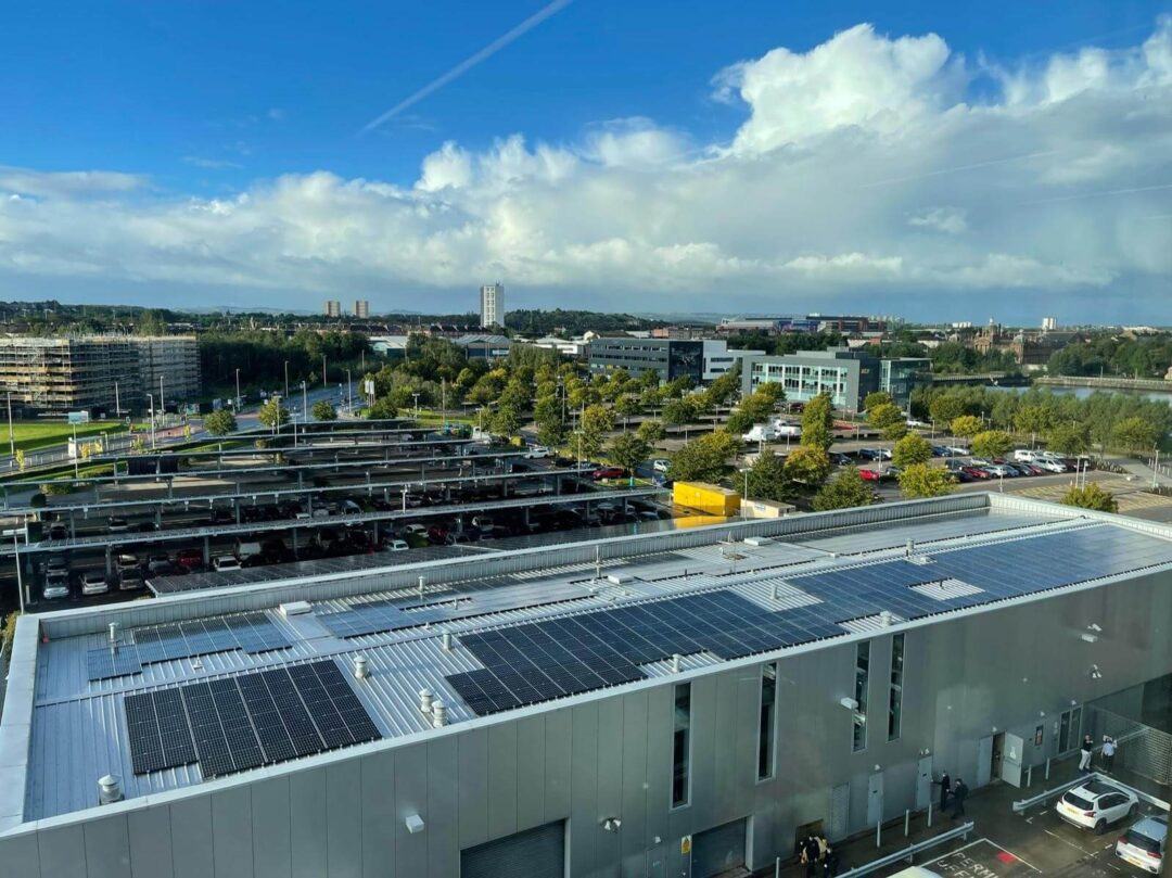 Aerial view of a commercial building with rooftop solar panels, overlooking a large multi-storey car park and surrounding business district under a bright, partly cloudy sky.