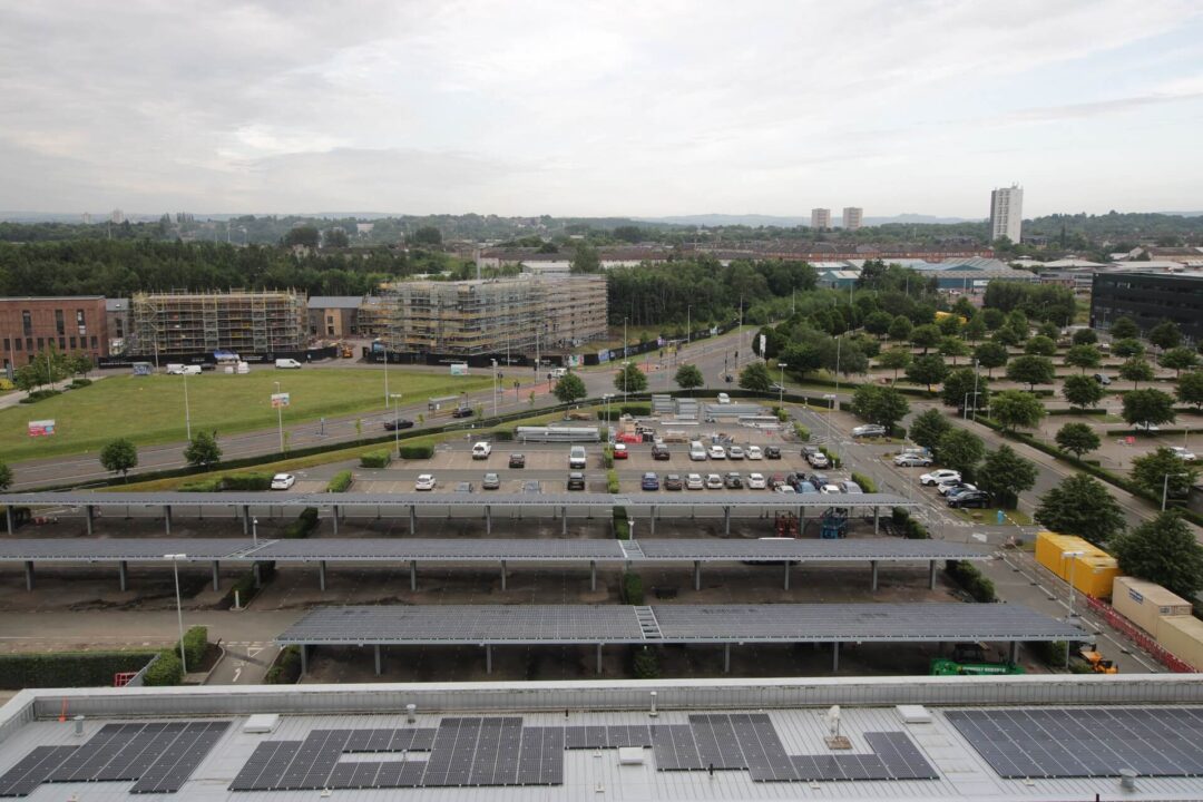 Aerial view of solar carports and rooftop solar panels at a commercial site, with surrounding parking areas, new building construction, and tree-lined roads in the background.