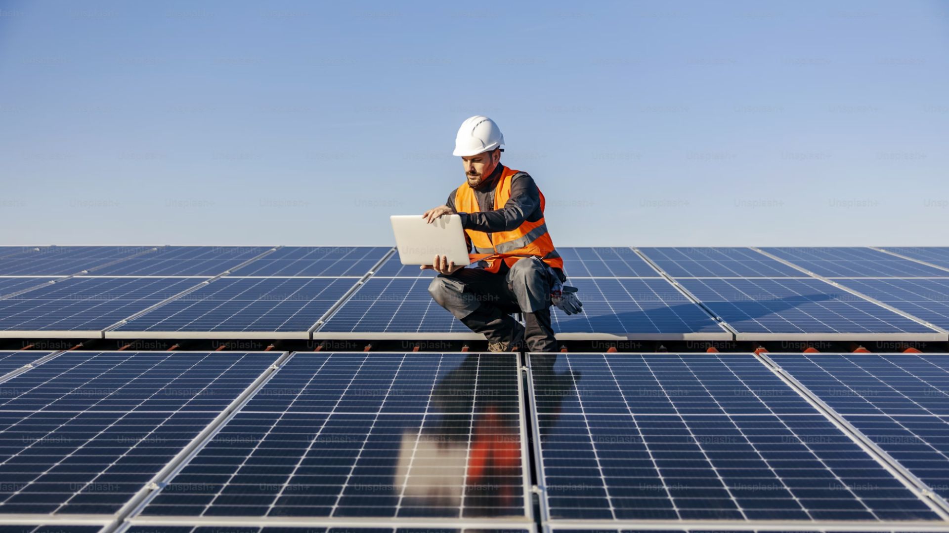 A man in an orange vest and hard hat is seated on a solar panel, engaged in work related to solar energy installation