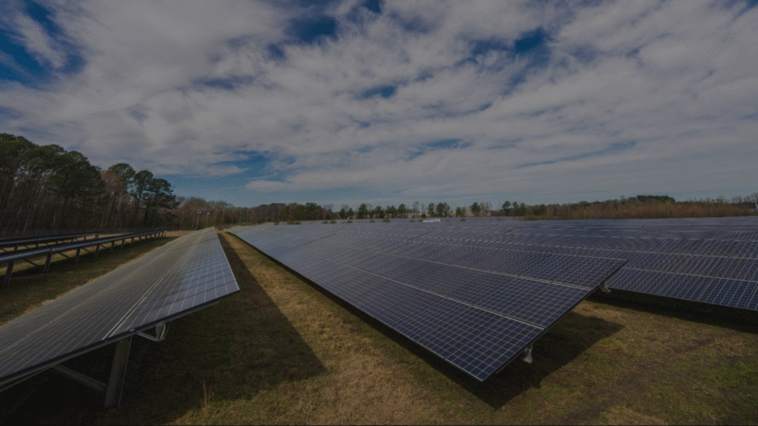 Wide ground-mounted solar farm with long rows of photovoltaic panels stretching across a grassy field under a partly cloudy blue sky, bordered by trees in the distance.