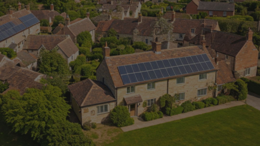 Aerial view of a traditional stone village house with a long row of solar panels on its tiled roof, surrounded by mature trees, gardens, and neighbouring cottages.