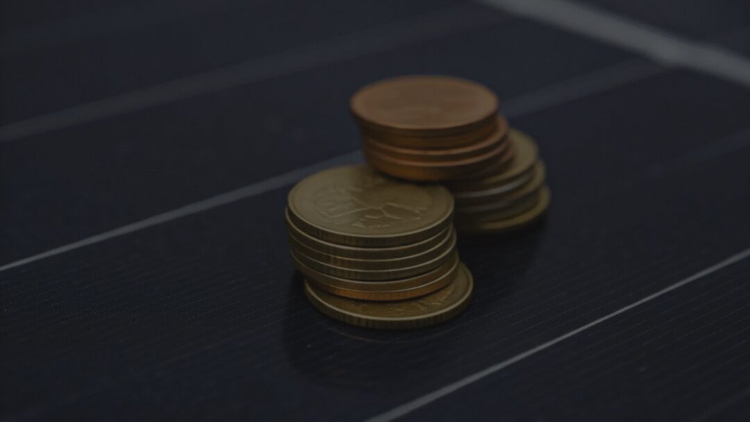 A close-up shot of stacks of copper and gold-colored coins resting on top of a dark, textured solar panel surface with visible white grid lines.