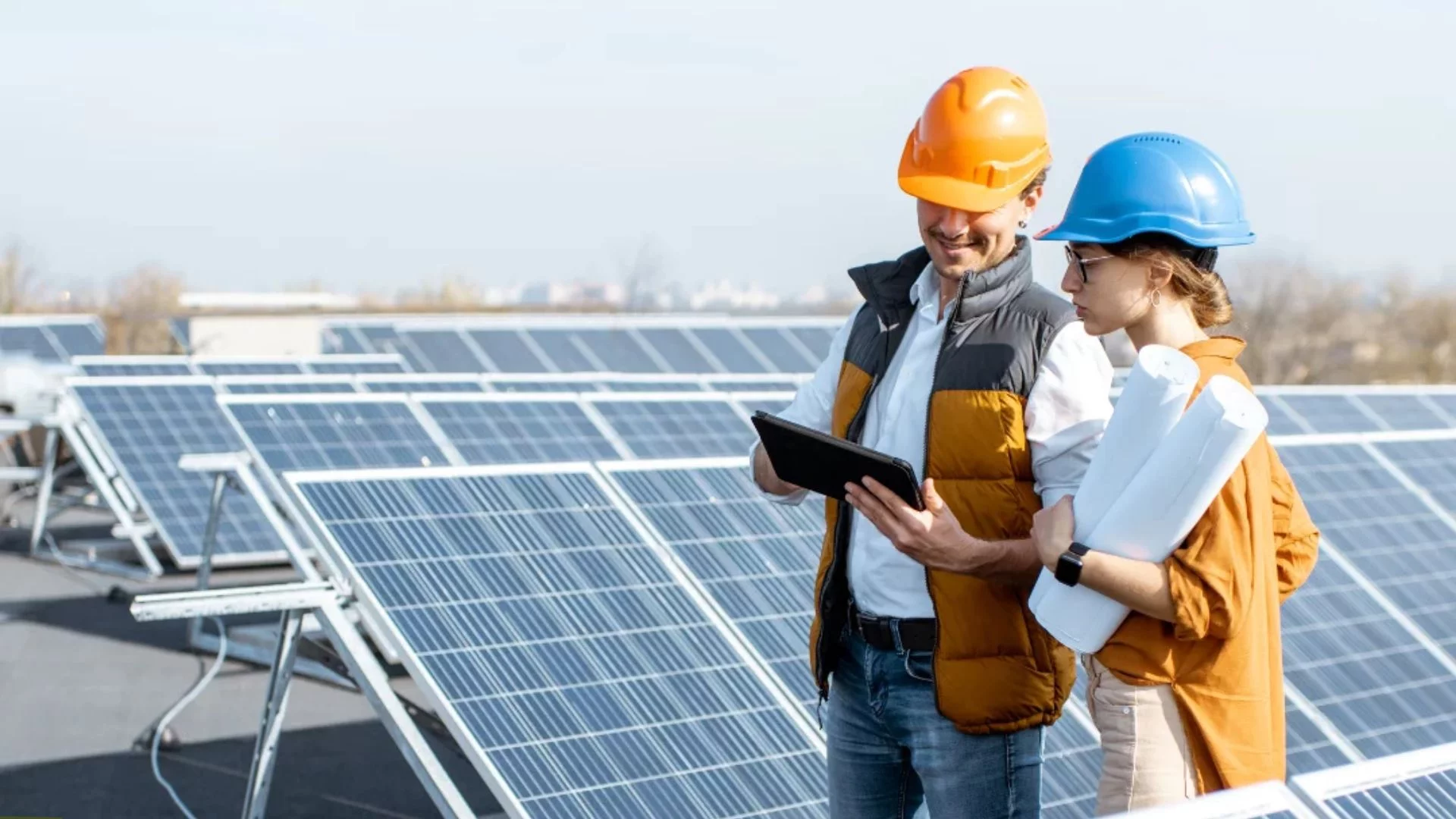 Two workers wearing hard hats are positioned next to solar panels, highlighting their involvement in solar energy projects.