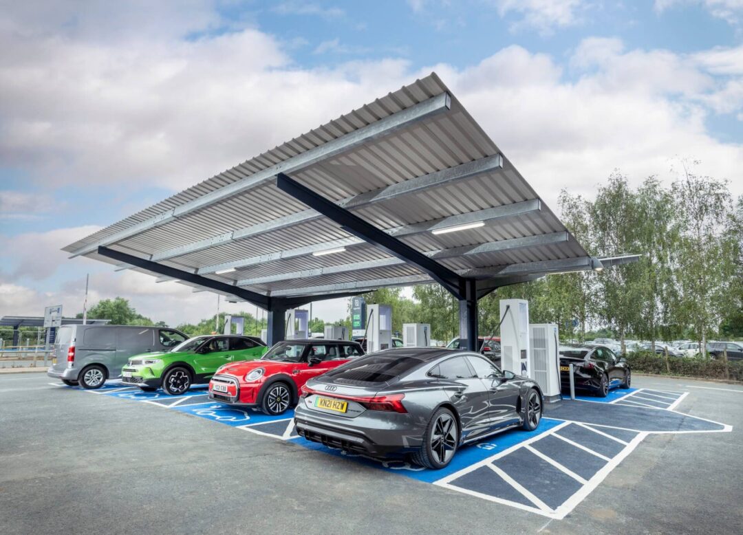 Covered electric vehicle (EV) charging station canopy with modern fast charging units, showcasing various electric cars like an Audi e-tron GT and a Mini in Solar Carports