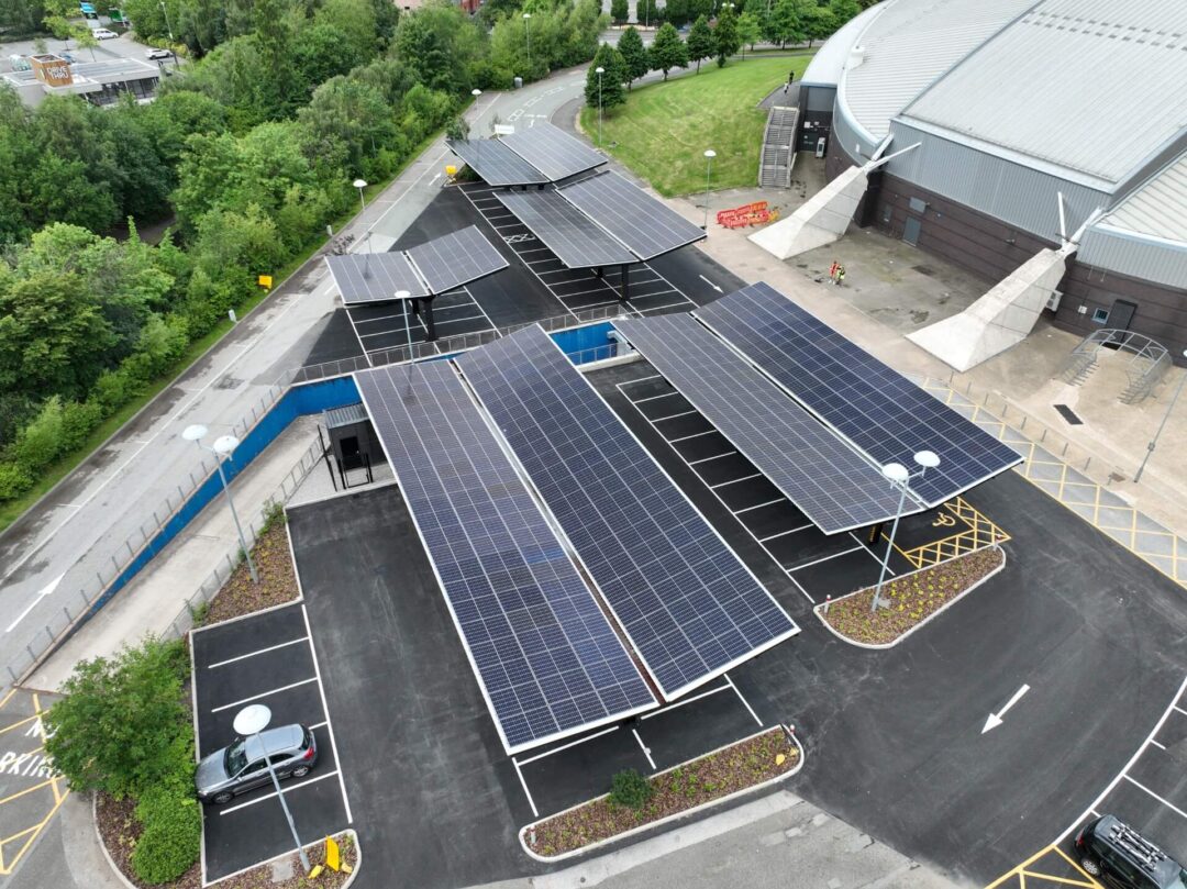 Aerial view of a multi-section solar panel carport installation over a large parking lot adjacent to a sports arena or public facility in Solar Carports