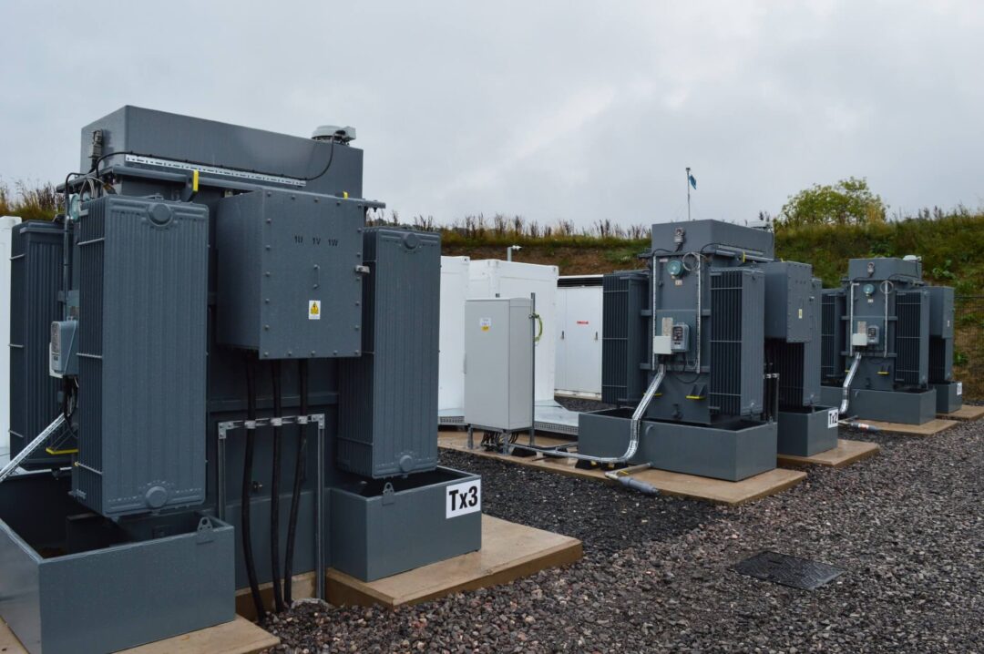 Ground level view of multiple large grey industrial transformers (Tx3) and white battery energy storage system (BESS) containers on a gravel pad at a substation in HV/LV Infrastructure