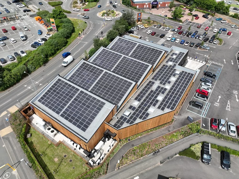 Aerial view of a commercial building with extensive rooftop solar panels, surrounded by busy roads and parking areas in a retail district in Reducing your company Carbon Footprint