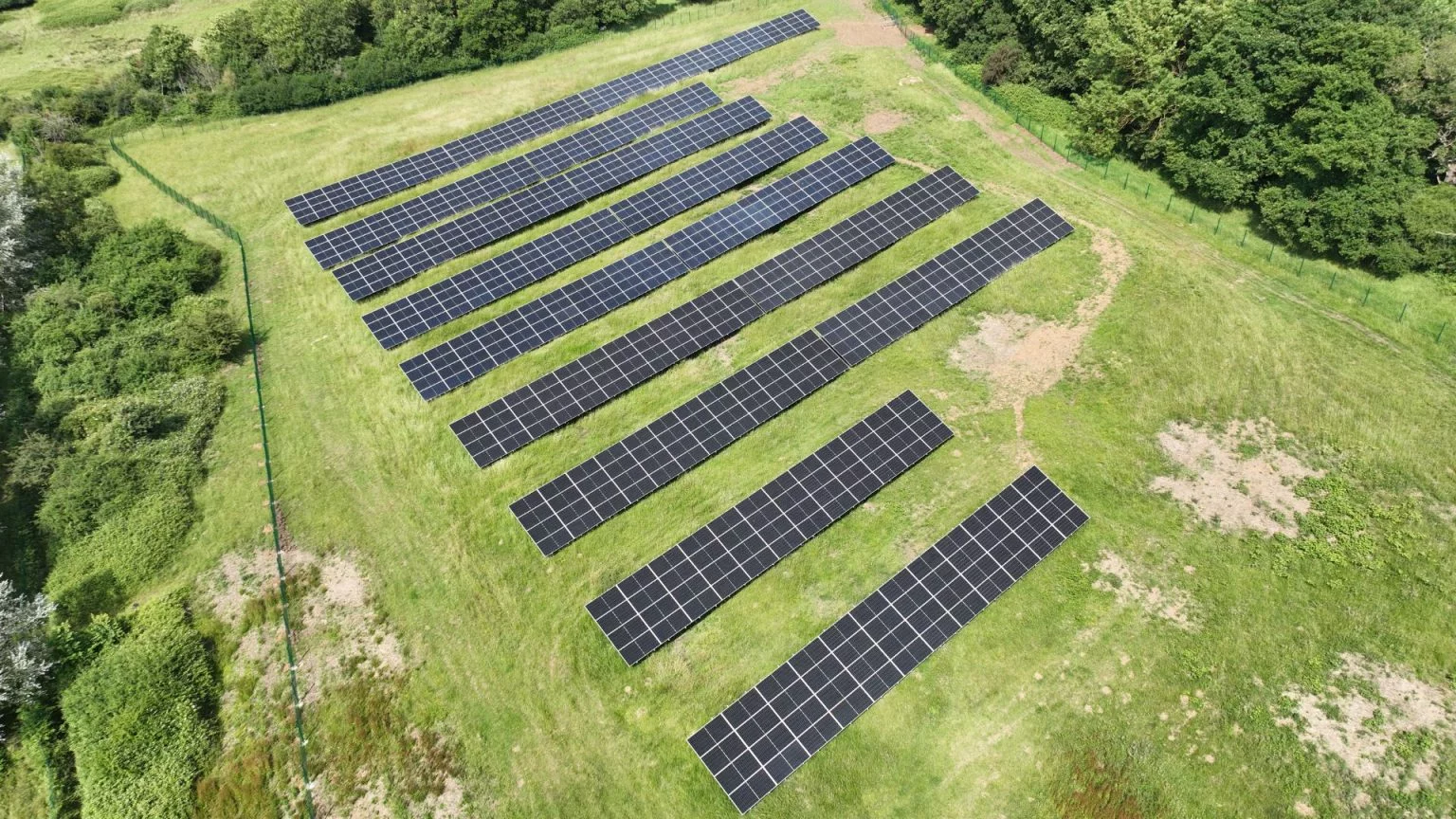 Aerial view of a small ground-mount solar PV installation with multiple rows of solar panels arranged on open grassland surrounded by trees.