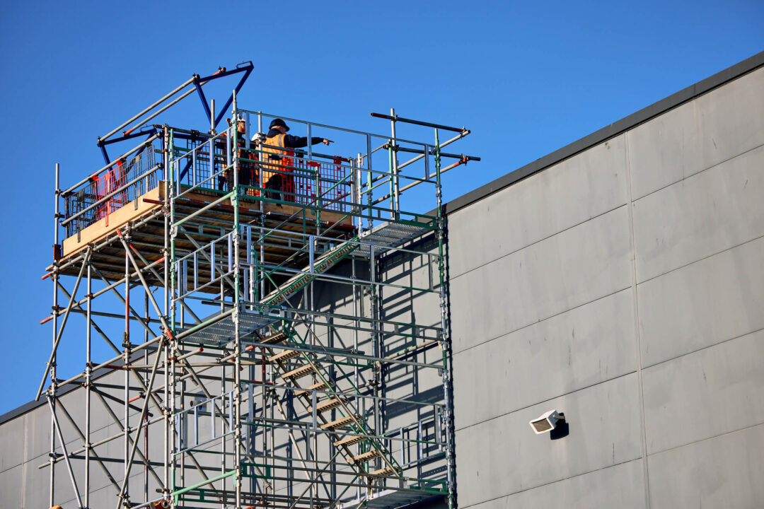 Engineers standing on a tall external scaffold structure beside a commercial building, preparing for rooftop installation work.