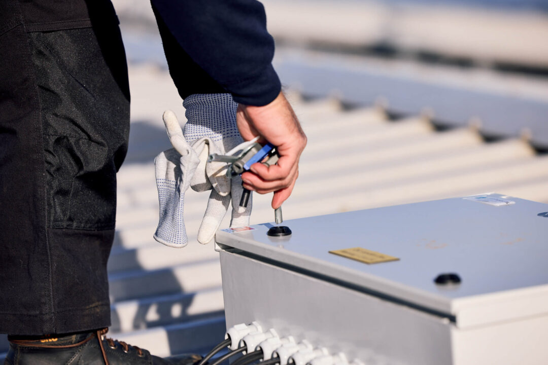 Engineer tightening a bolt on an electrical junction box during a rooftop solar installation, with tools and safety gloves visible.