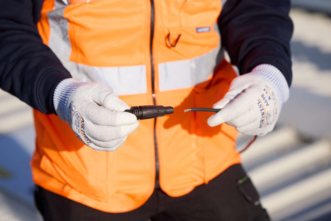 An engineer wearing safety gloves and a high-visibility vest preparing to connect solar PV wiring using an MC4 connector.