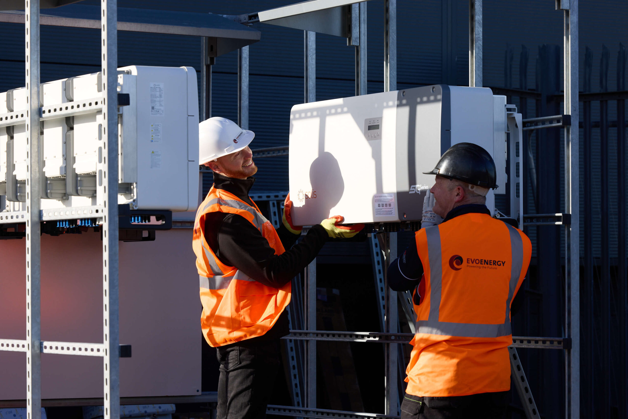 Two EvoEnergy engineers in safety gear lift and position a large solar inverter onto its mounting frame at an installation site.