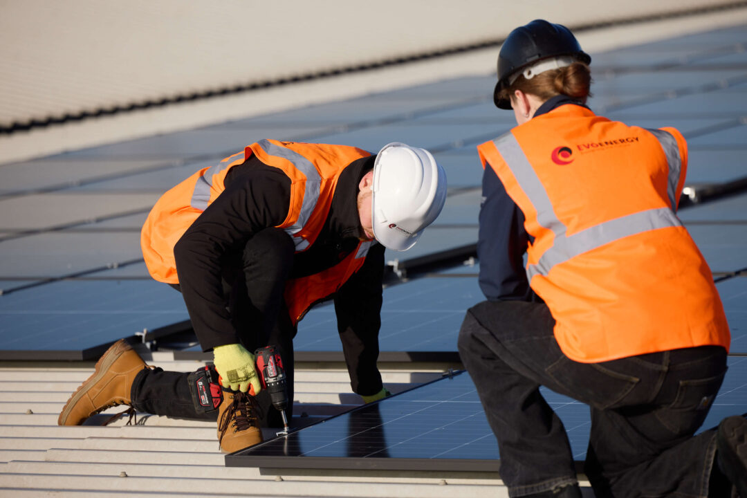Two engineers in high-visibility vests and hard hats installing solar panels on a commercial rooftop, using power tools to secure the mounting system.