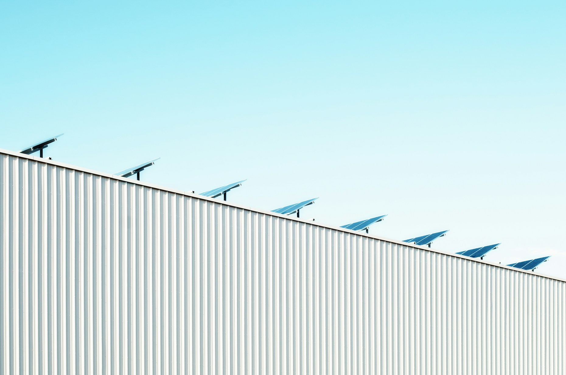 Minimalist view of solar panels installed along the edge of a large industrial metal roof against a clear blue sky.