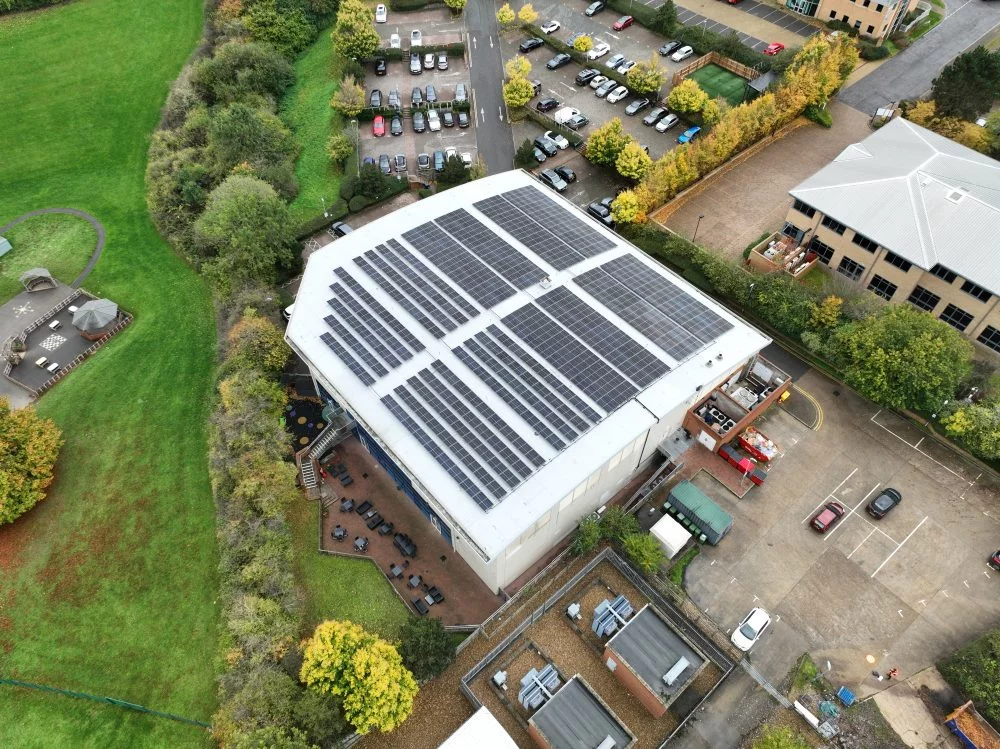 Aerial view of a commercial building with a large rooftop solar panel installation surrounded by green space and adjacent parking areas.
