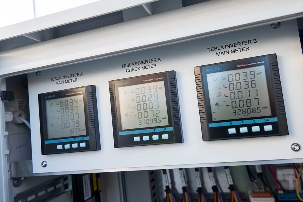 Close-up of Tesla inverter power meters displaying live solar generation and system performance data inside an electrical control panel in Solar Output Calculator