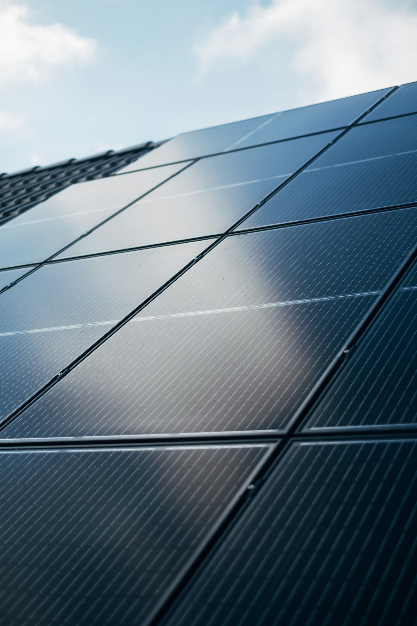 Close-up view of black solar panels installed on a sloped roof with a bright blue sky and clouds reflected on the panel surface.