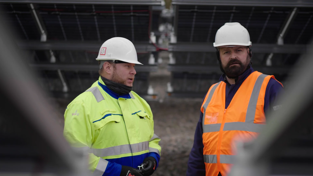 Two construction workers wearing hard hats and high-visibility jackets discussing in front of a solar panel installation site.