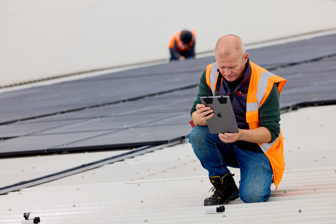 Solar engineer in high-visibility PPE inspecting a rooftop solar installation and reviewing data on a tablet during a commercial PV project.