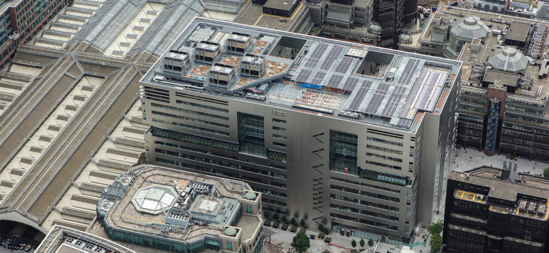 Aerial view of a modern office building with rooftop solar panels and mechanical plant equipment, surrounded by dense city architecture.