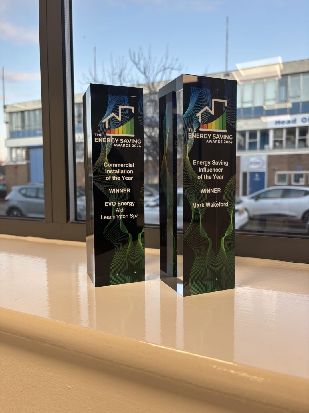 Two Energy Saving Awards 2024 trophies displayed on a windowsill. One award recognises EvoEnergy for Commercial Installation of the Year for the Aldi Leamington Spa project, and the other recognises Mark Wakeford as Energy Saving Influencer of the Year. A blurred office and parked cars are visible outside the window.