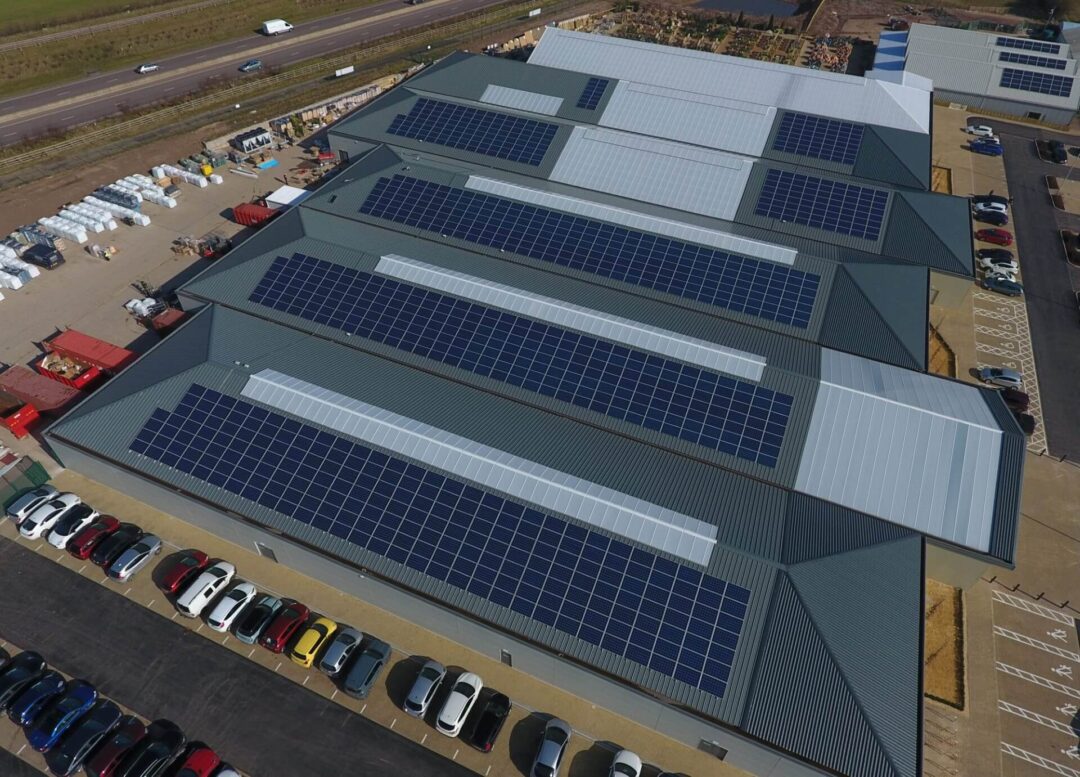 Aerial view of a large commercial warehouse with multiple rooftops covered in solar PV panels, with parked cars lining the building.