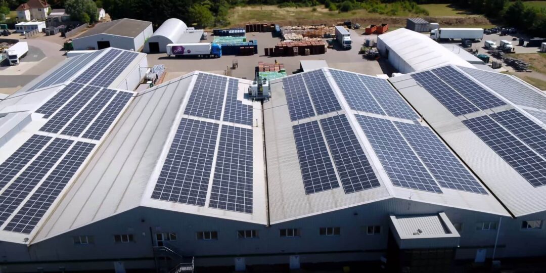 Aerial view of a large industrial warehouse roof covered with multiple rows of solar PV panels, with storage yards, trucks, and surrounding buildings visible in the background.