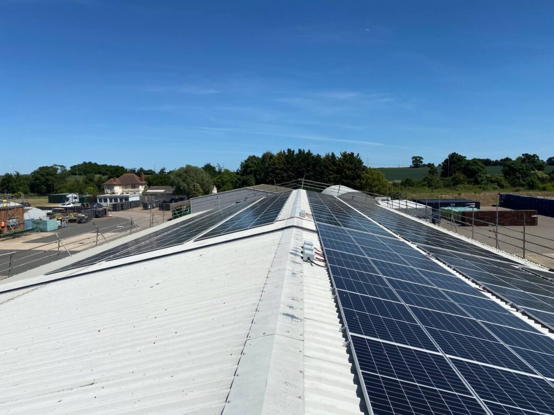 Rooftop solar panel installation on a large industrial building, shown under a clear blue sky with surrounding warehouses, fields, and rural landscape in the background.