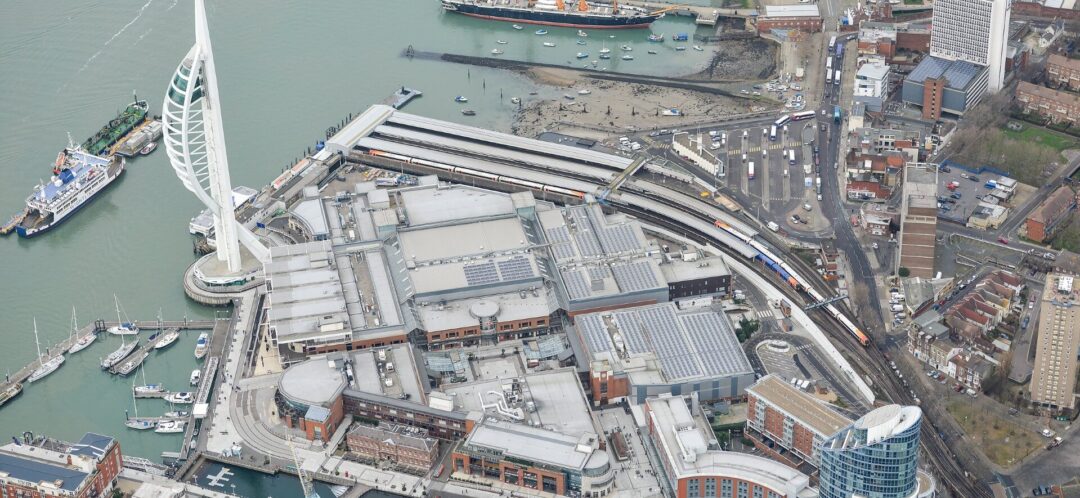 Aerial view of Portsmouth Harbour showing the Spinnaker Tower, waterfront buildings with rooftop solar panels, marina boats, and nearby railway tracks with trains.