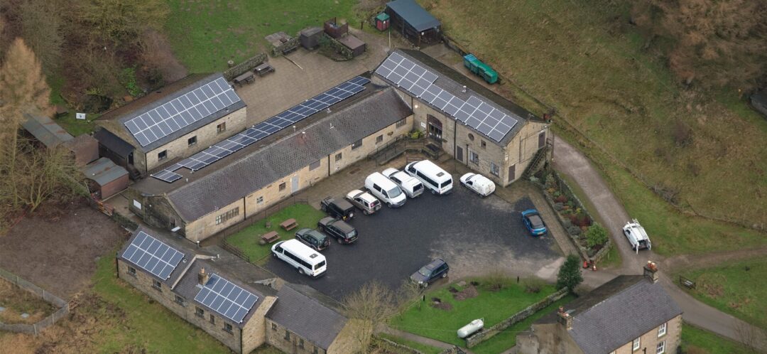 Aerial view of a rural courtyard complex with stone buildings fitted with rooftop solar panels and several parked vehicles.