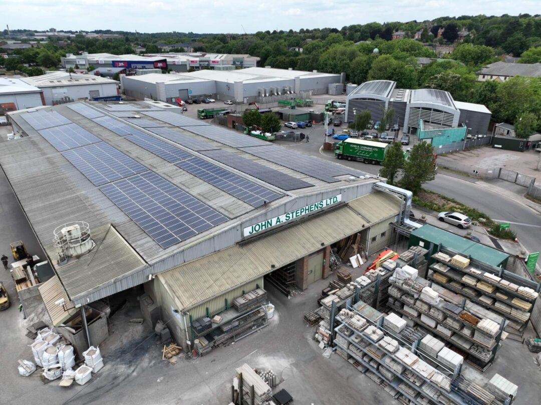 Industrial warehouse at John A. Stephens Ltd with a large rooftop solar PV installation, surrounded by storage racks, construction materials, and nearby commercial buildings.