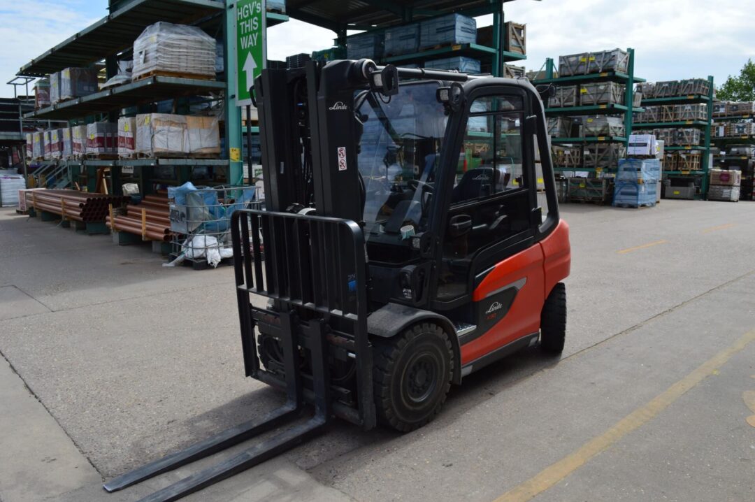 A red and black Linde forklift parked in an outdoor industrial yard, surrounded by stacked building materials and storage racks in John A Stephen