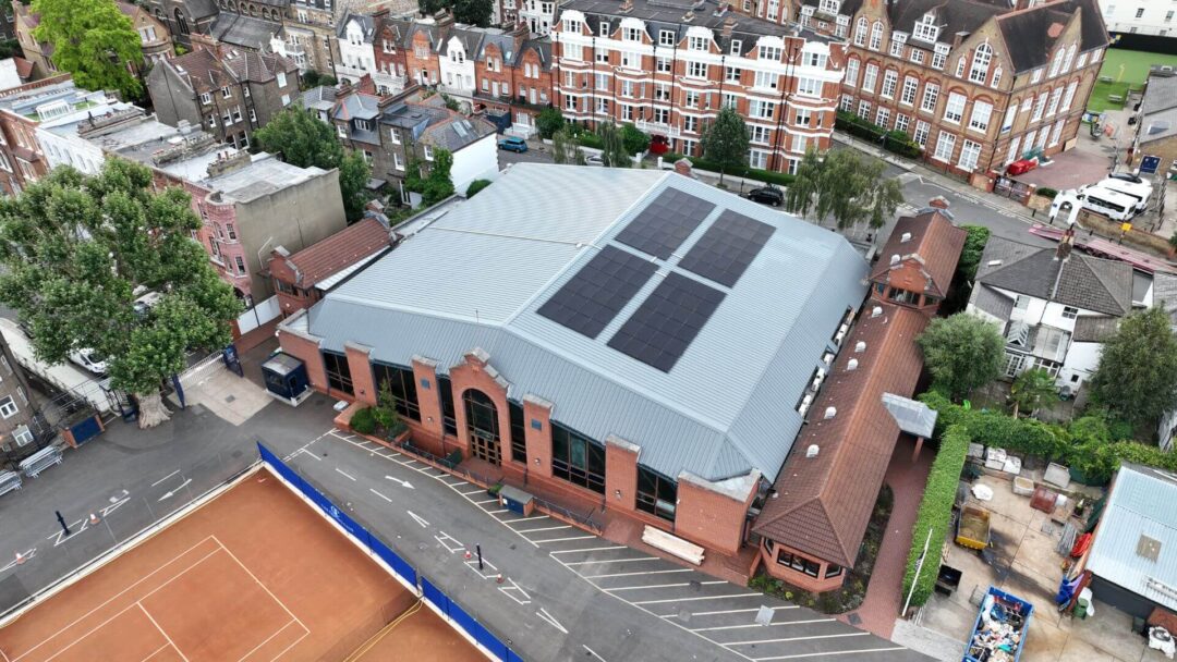 Aerial view of a commercial sports facility with rooftop solar panels installed on a large grey metal roof, surrounded by residential buildings and outdoor tennis courts in Queens Club.
