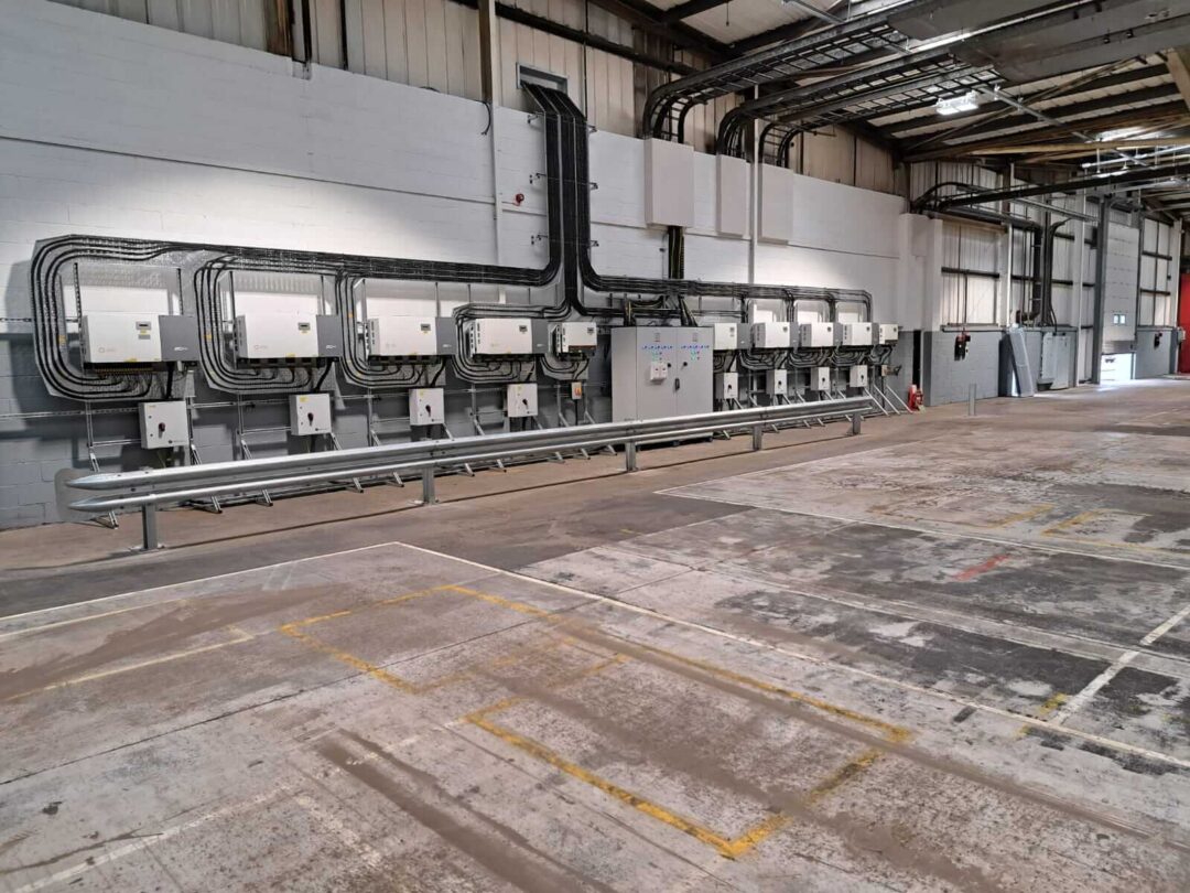 Interior view of an industrial facility showing multiple solar PV inverters neatly mounted on a wall with organised cabling and protective barriers in front in Rainhill Road