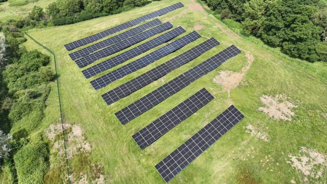 Aerial view of a small ground-mounted solar PV array installed on a grassy field, bordered by trees and fencing, used for renewable energy generation in Scouts