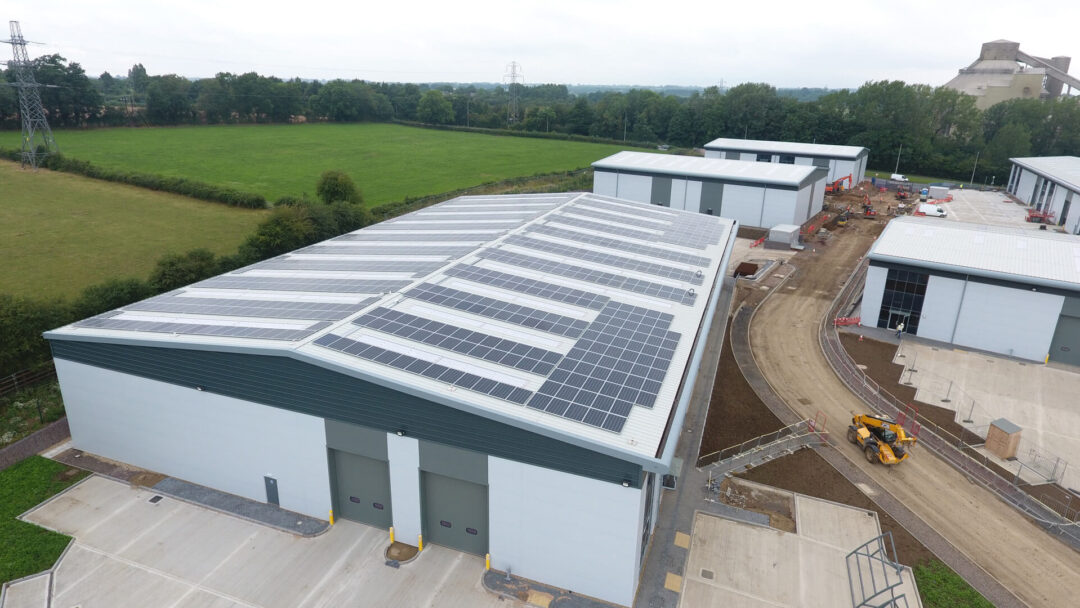 Drone view of a commercial industrial estate with solar panels installed across the roof of a large warehouse building, surrounded by green fields and ongoing construction works.
