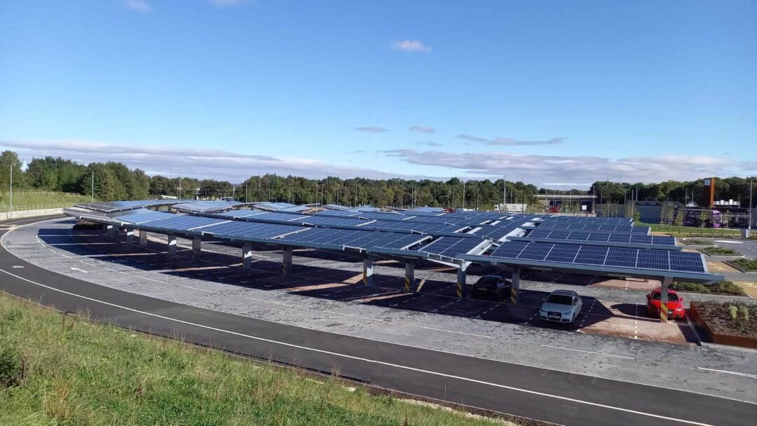 Solar carport structures covering parking bays at a modern car park, with several vehicles parked beneath and trees visible in the background.