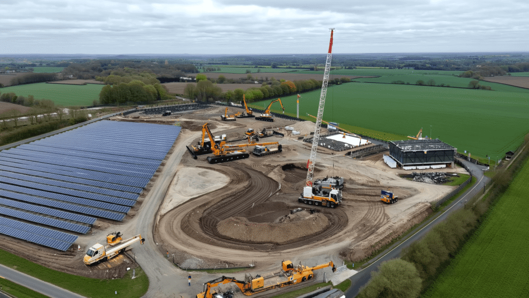 Aerial view of a large solar farm under construction, featuring installed solar panel arrays on one side and multiple cranes and heavy machinery working on site development across a rural landscape.
